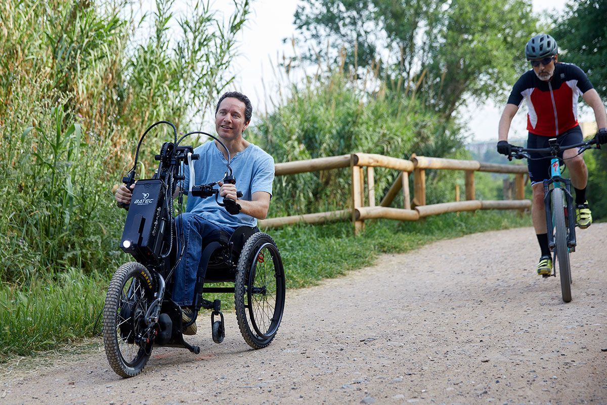 Man in wheelchair bike on gravel path, another cycles past. Outdoors.