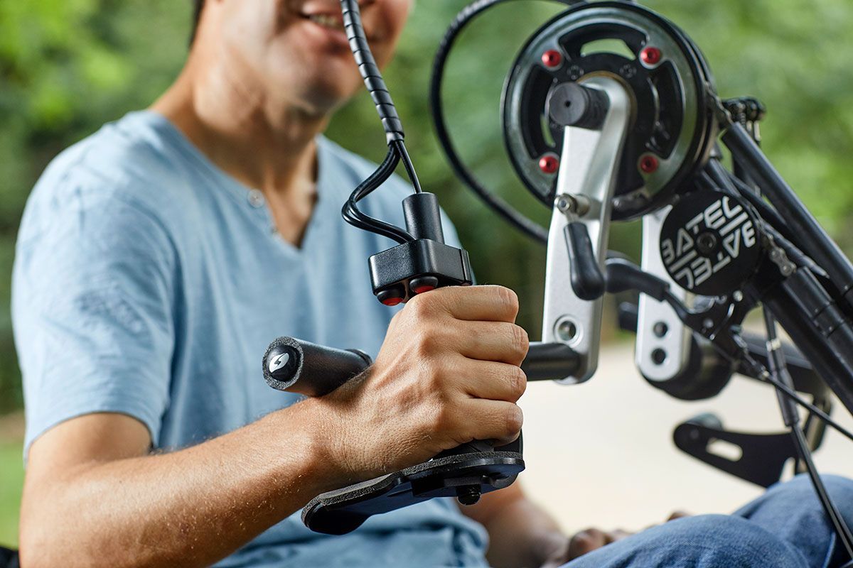 Man operating handcycle outdoors, smiling. Black handgrip, mechanical details visible.