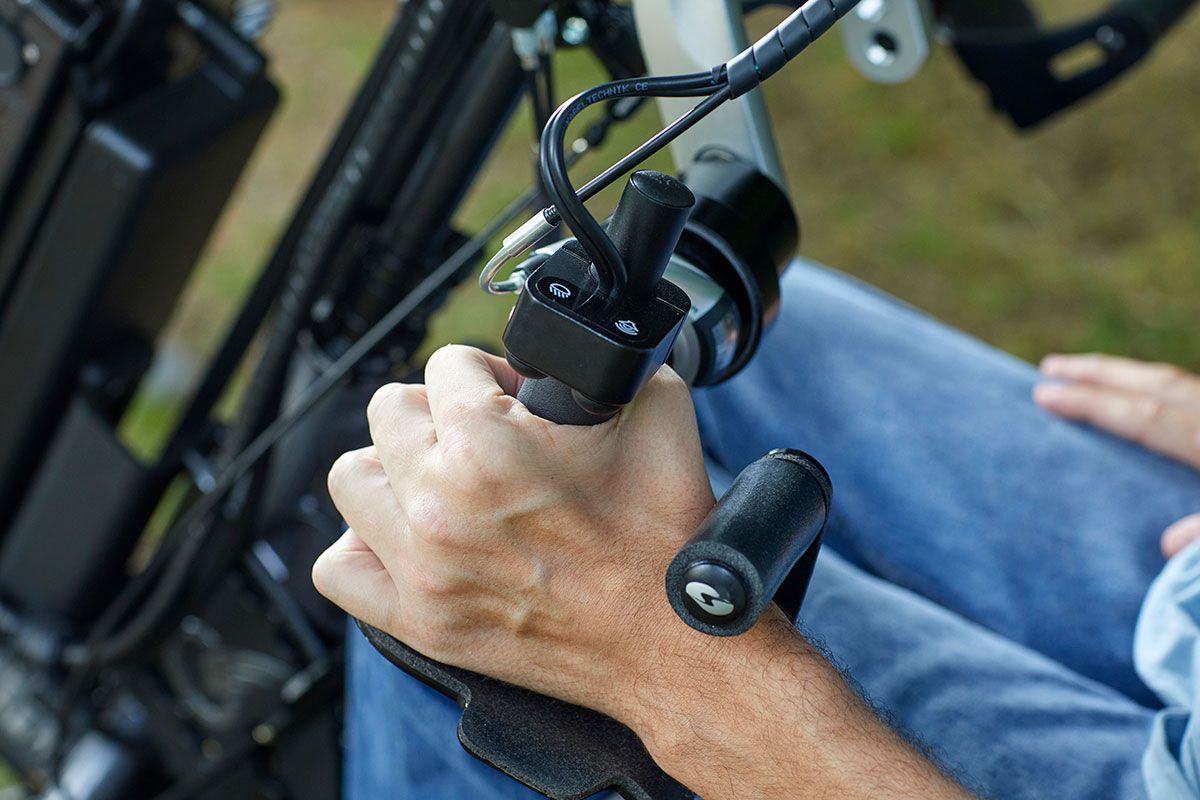 Person's hand on a power wheelchair joystick. The hand rests on the black control, and the background is blurred.