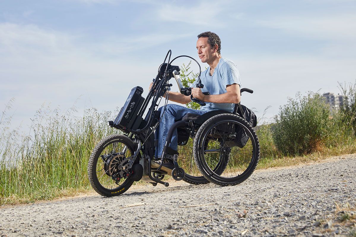 Man using a black all-terrain wheelchair on a gravel path outdoors. Sunny day.