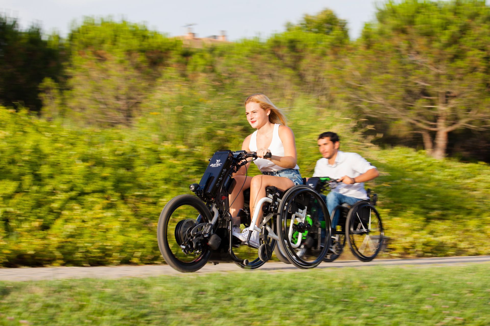 Woman and man in wheelchairs ride on a path. The woman's chair has an electric motor. Outdoors, sunny day.