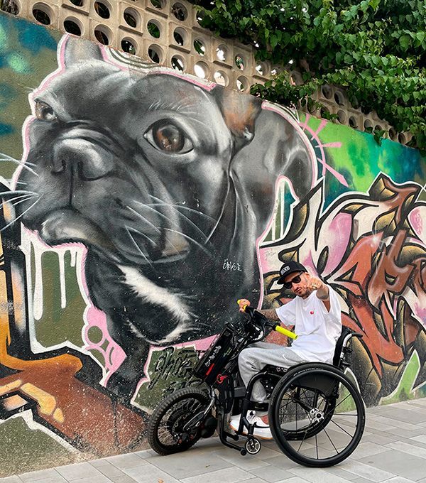 Man in wheelchair in front of a bulldog mural, gesturing with his hand.