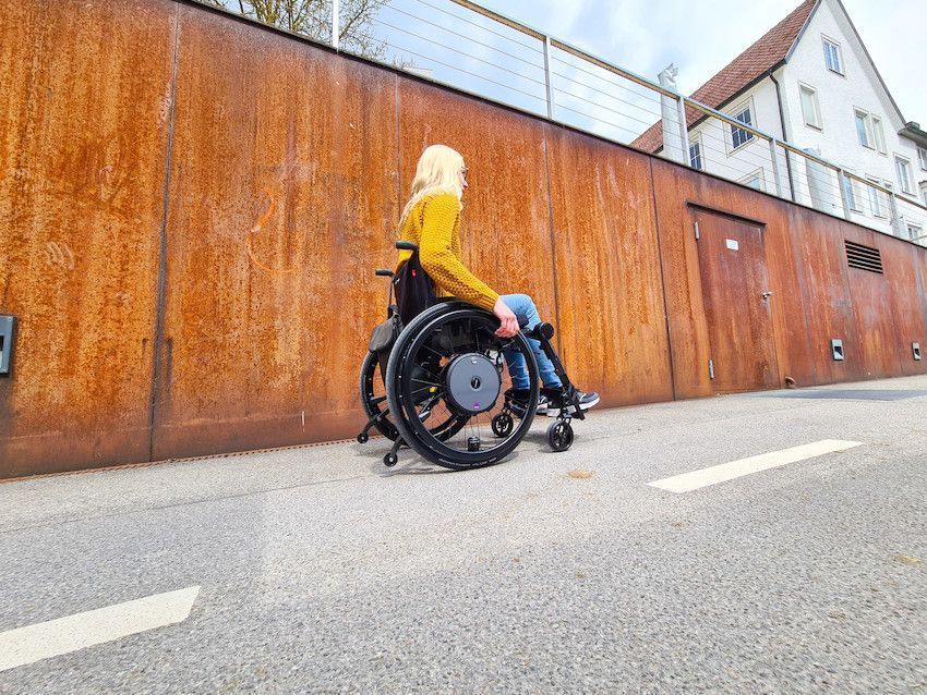 Woman in wheelchair on a paved path next to a rusty metal wall and a building. Sunny day.
