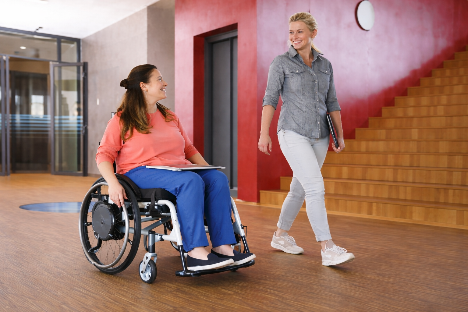 Woman in wheelchair smiling, talking to woman walking with tablet; indoor setting with stairs and red wall.