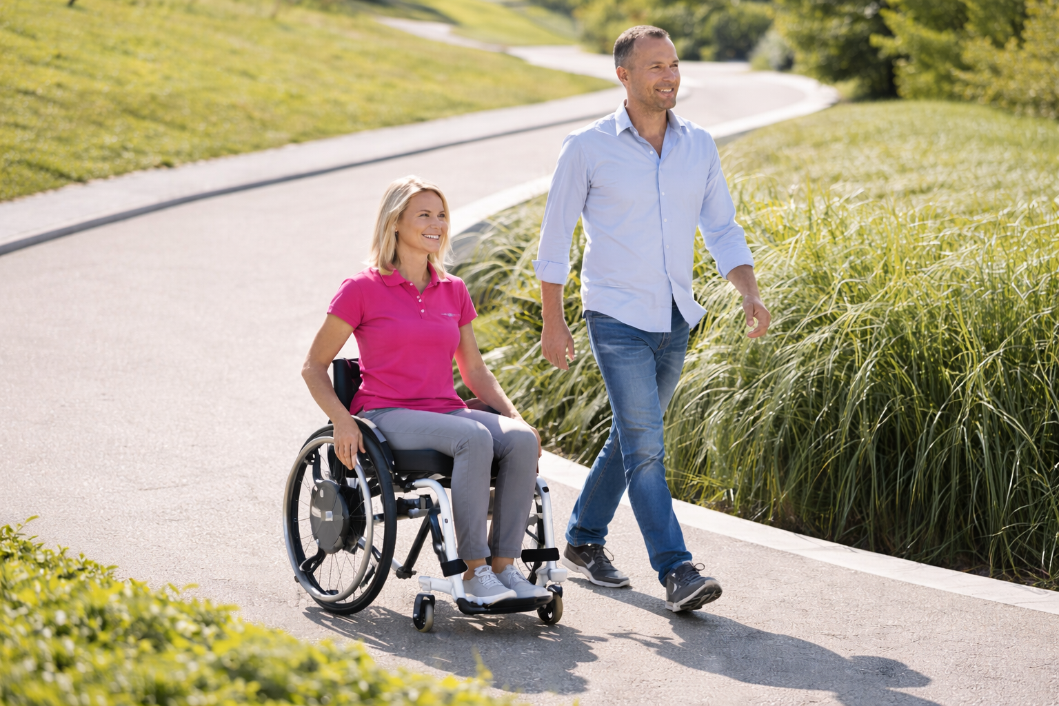 Woman in wheelchair and man walk on a paved path in a park; sunny day.