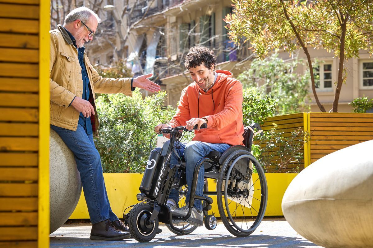 Man in wheelchair with another man, outdoors, smiles, sunshine.