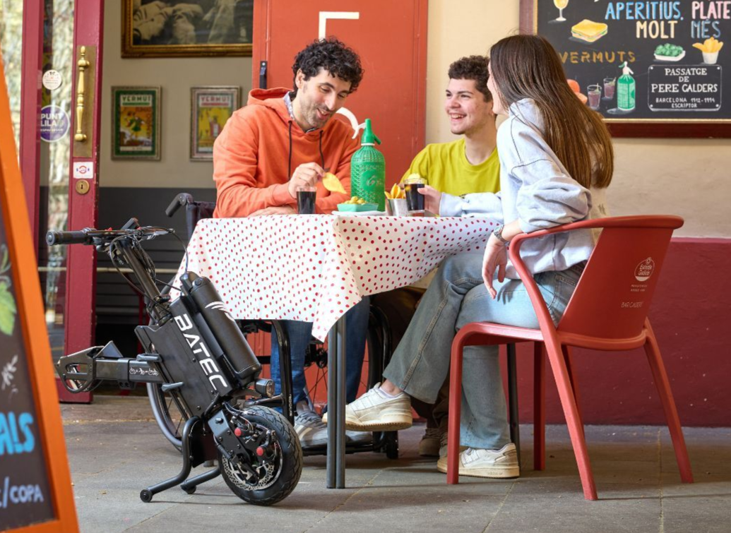 People at an outdoor cafe table with a BATEC electric bike parked nearby; enjoying drinks and conversation.