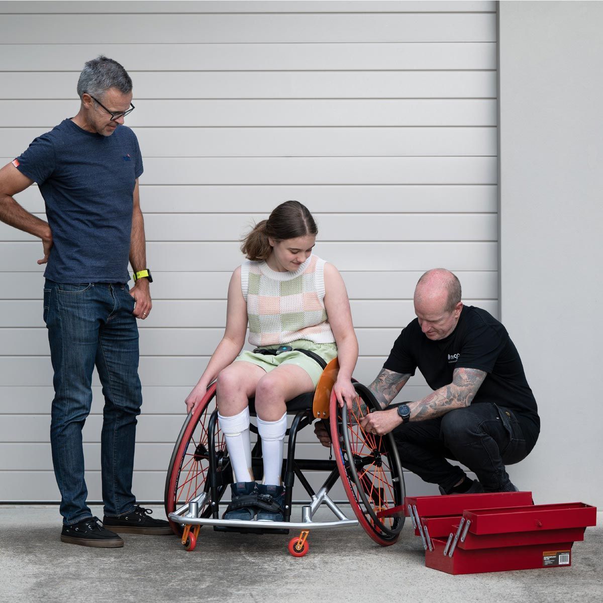 Three people with a wheelchair, one adjusting the wheel, the other observing. Gray garage door background.