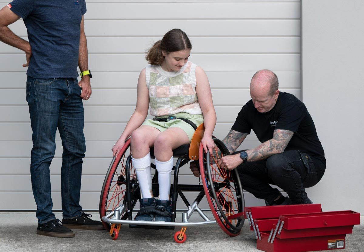 A person in a wheelchair having their wheel adjusted by a person while another person watches; red toolbox nearby.