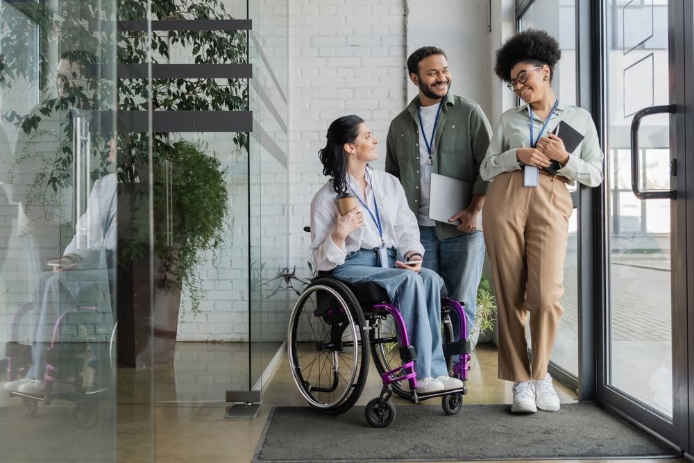 Three office workers, one in a wheelchair, smiling and talking by a glass door.