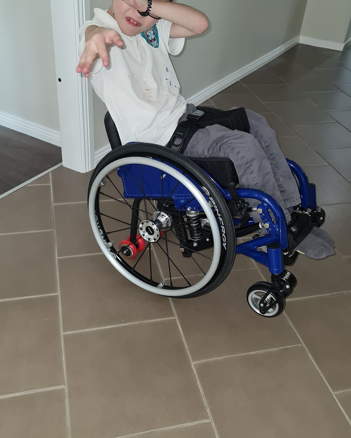 Boy in wheelchair poses with a hand gesture, indoors on tile flooring. Blue wheelchair, white shirt.