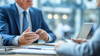 Business meeting: Man in blue suit gestures, discussing documents at a table.