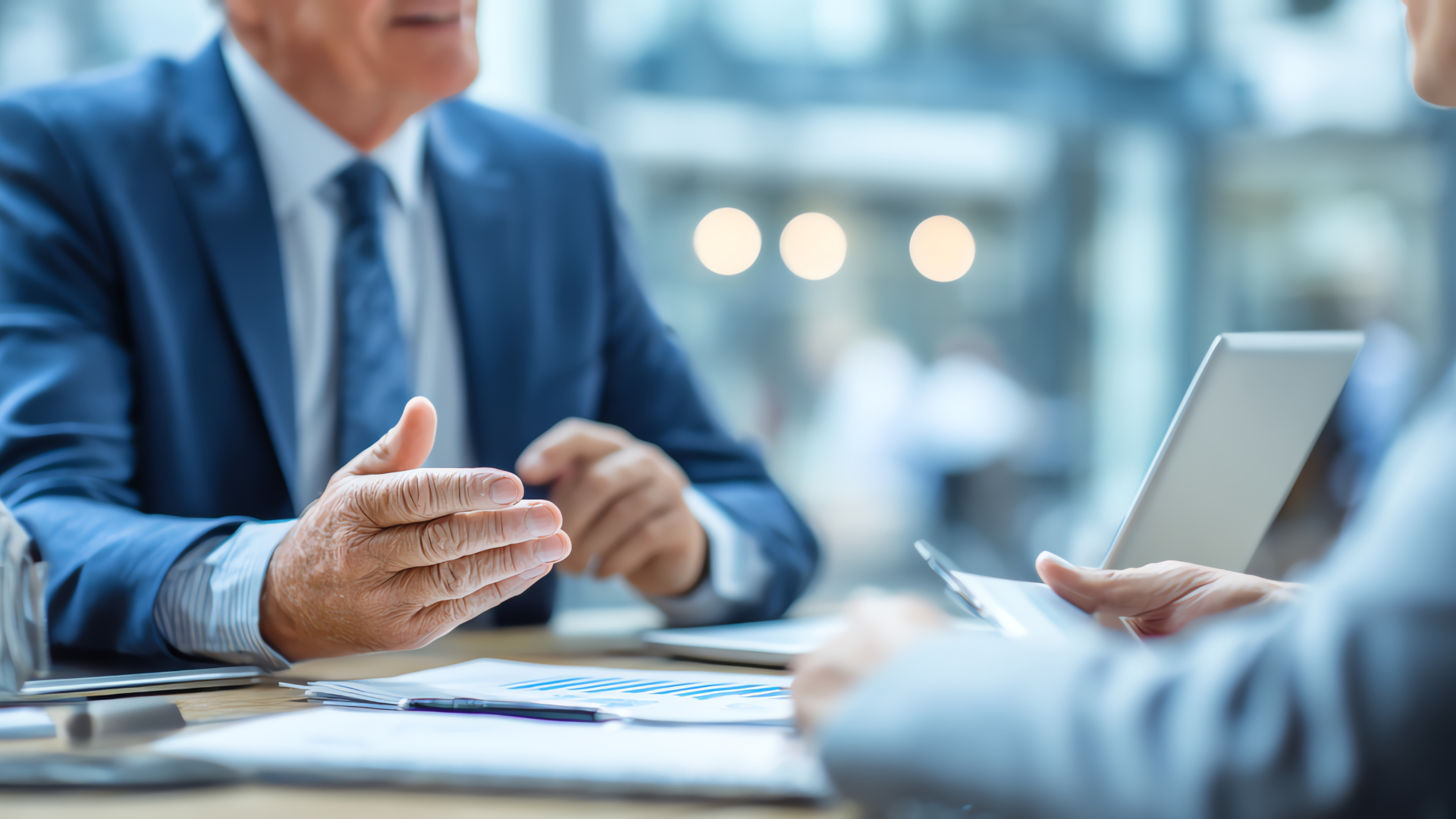 Business meeting: Man in blue suit gestures, discussing documents at a table.