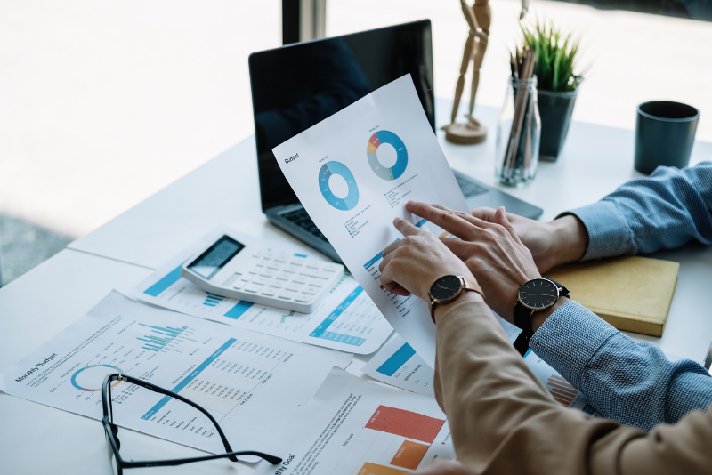 Hands pointing at financial charts with laptop, calculator, and coffee mug on a white desk.