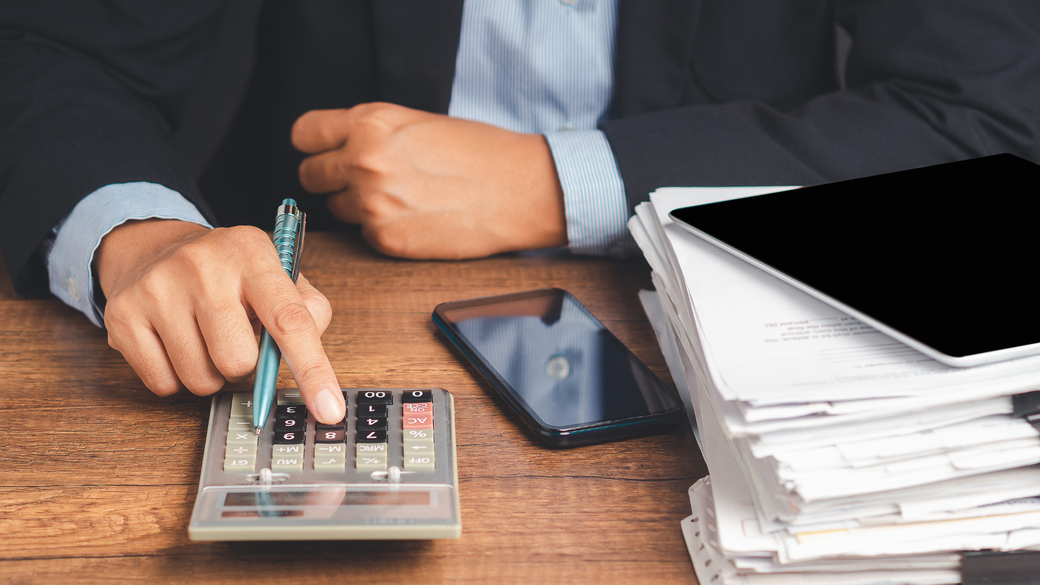 Person in suit calculating with calculator, papers, phone, and tablet on a wooden desk.