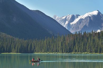 Canoe with two people on a clear turquoise lake, mountains and evergreen trees in the background.