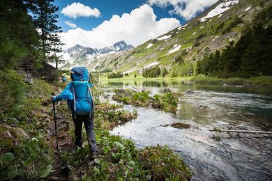 Hiker with blue backpack treks along a lush, streamside trail in a mountainous landscape.