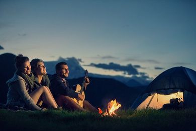 People around a campfire at dusk, mountains in background. One person plays guitar, tent nearby.