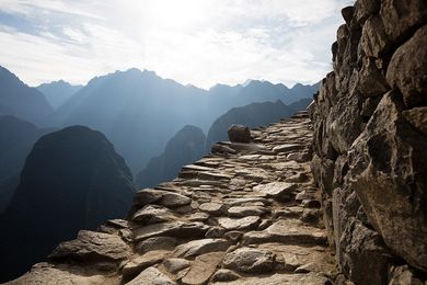 Stone path ascending a mountain, with a stone wall on the right and mountain range in the background under a bright sky.