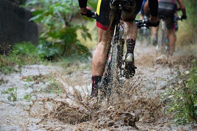 Cyclist riding through muddy water, legs and bike splattered, following other cyclists in a forest.