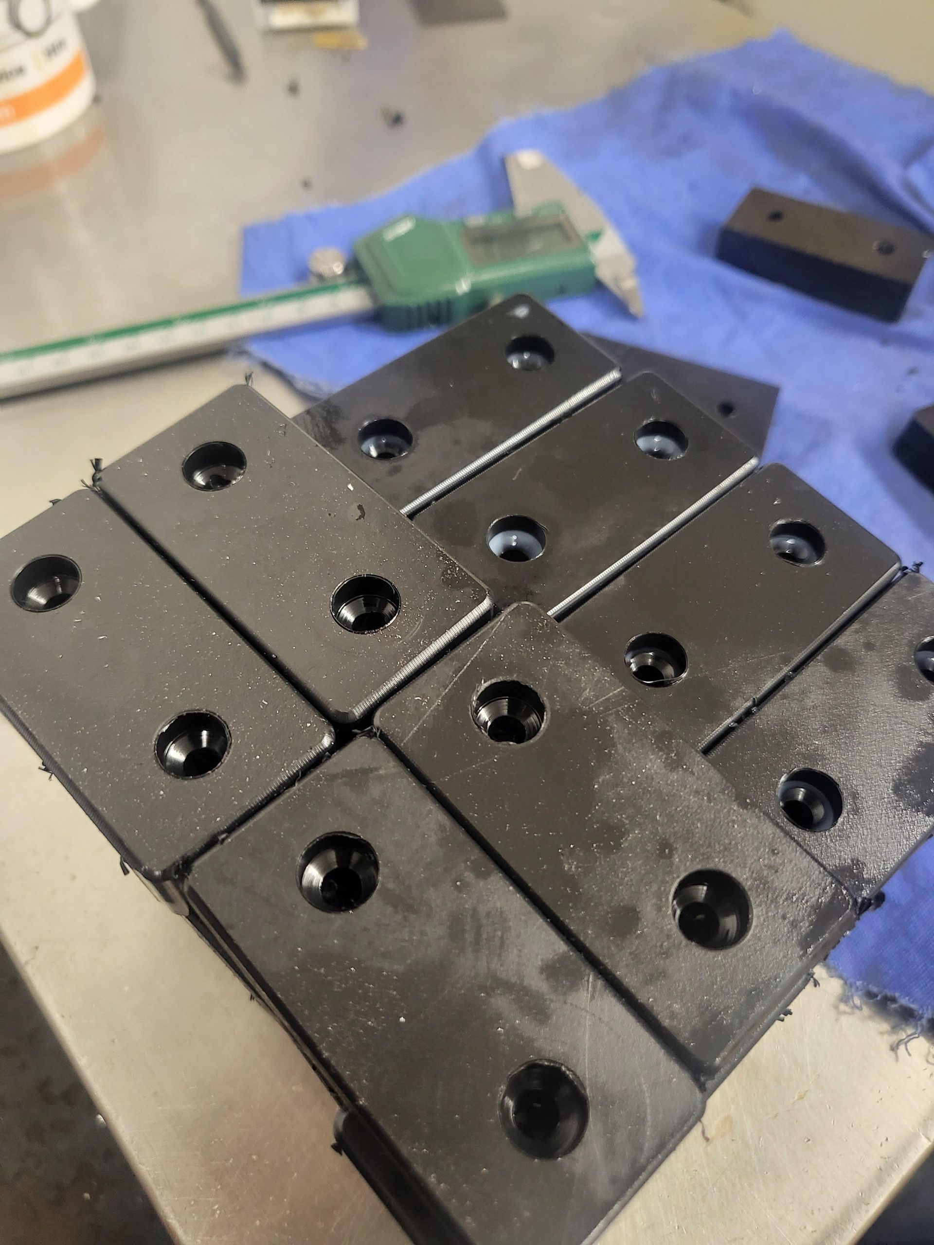 Black rectangular metal pieces with drilled holes, arranged in a grid, next to a caliper and blue cloth.