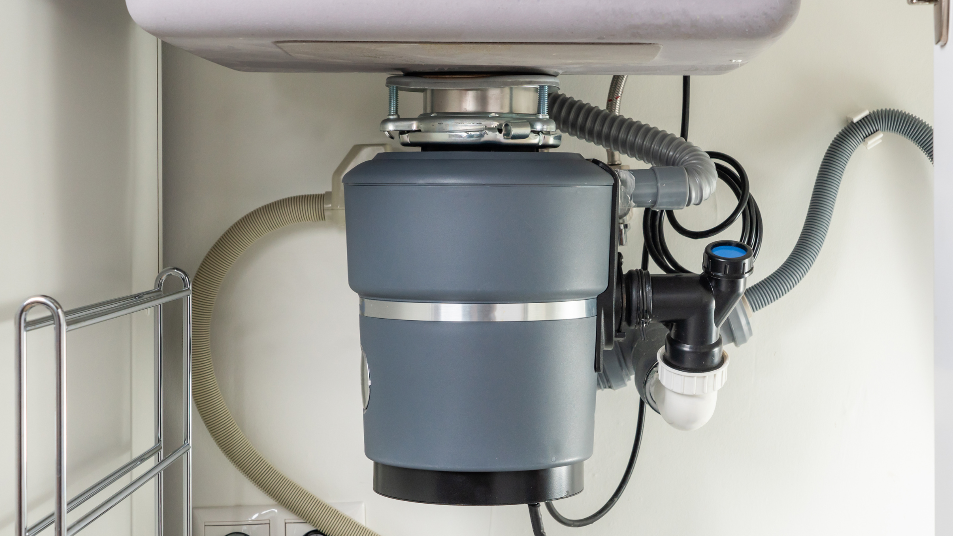 Garbage disposal unit under a kitchen sink, connected to plumbing and electrical wiring. Gray and white.