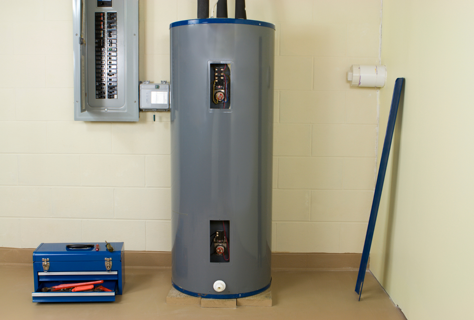A tall, gray water heater in a room with a circuit breaker box, toolbox, and level leaning against a wall.