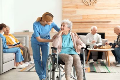 Caregiver helping a seated woman in a wheelchair in a bright nursing home common room