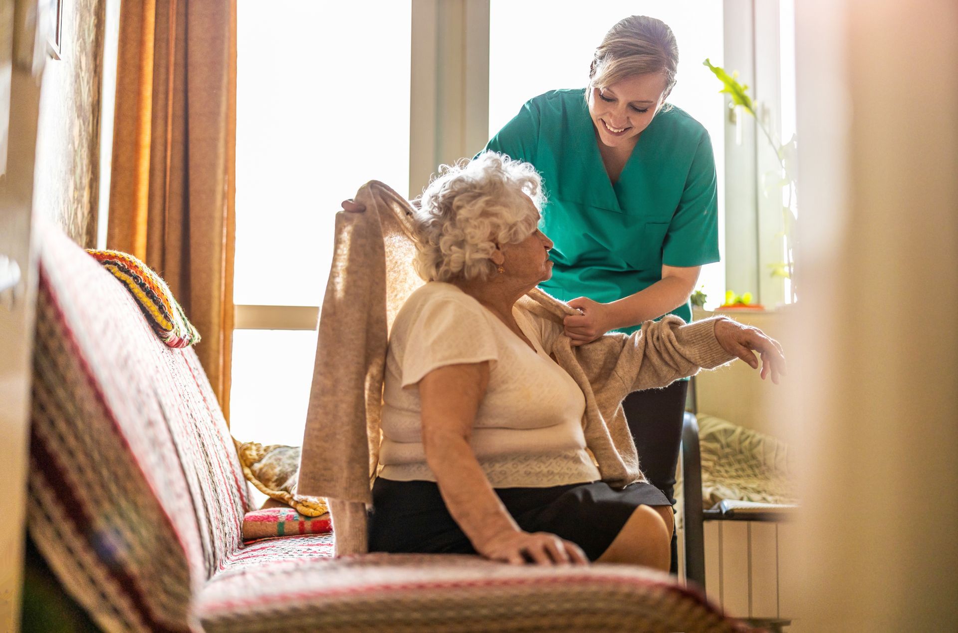 Caregiver in green helping a seated woman with a walker in a sunlit living room Caregiver in green helping a seated woman with a walker in a sunlit living room