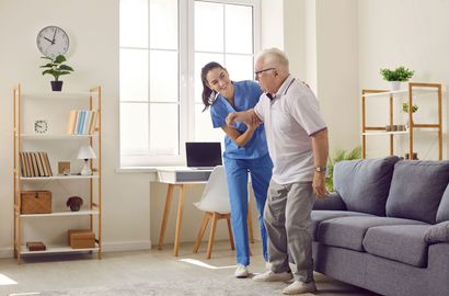 Older man and woman stretching in a bright living room beside a gray sofa and laptop table