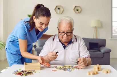 Caregiver in blue helps an elderly man sort colorful pieces at a table in a living room