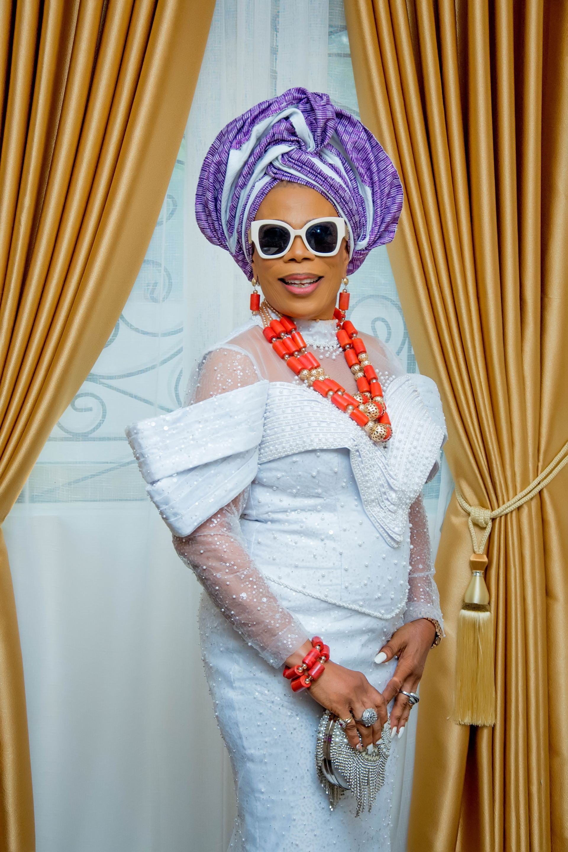 A woman in a white dress and purple turban is standing in front of a window.