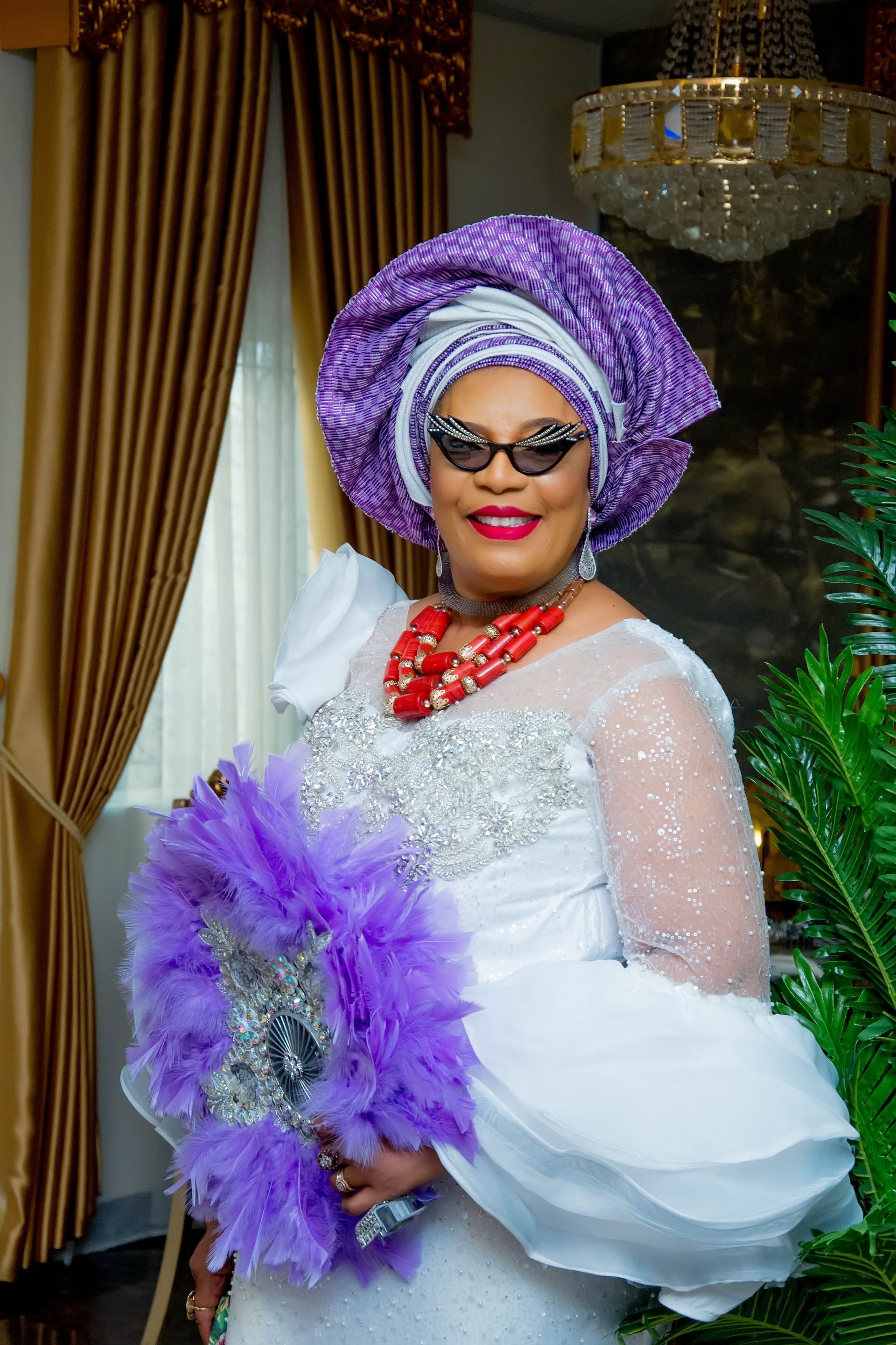A woman in a white dress and purple turban is holding a purple fan.
