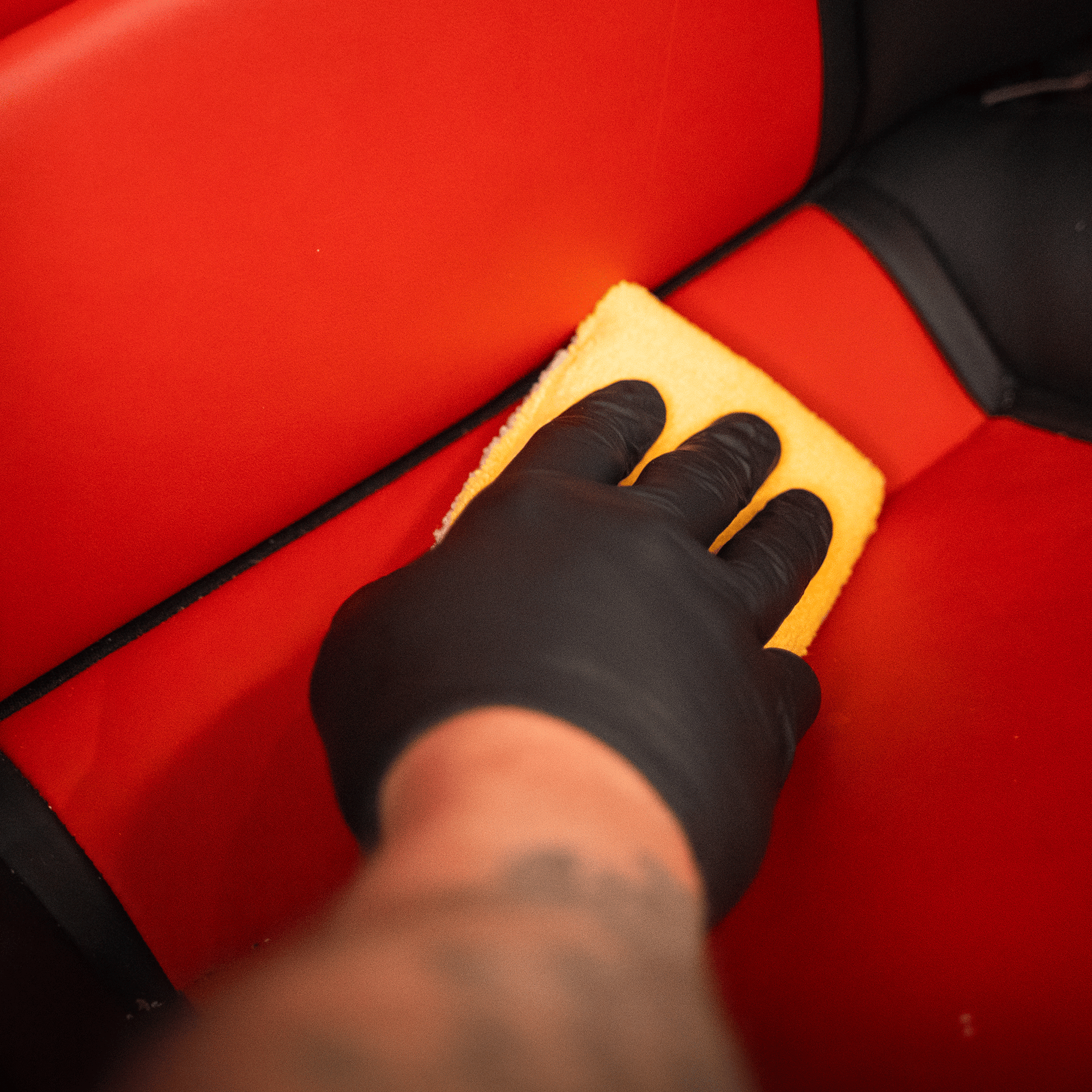 Gloved hand cleaning a bright red car seat with a yellow sponge.