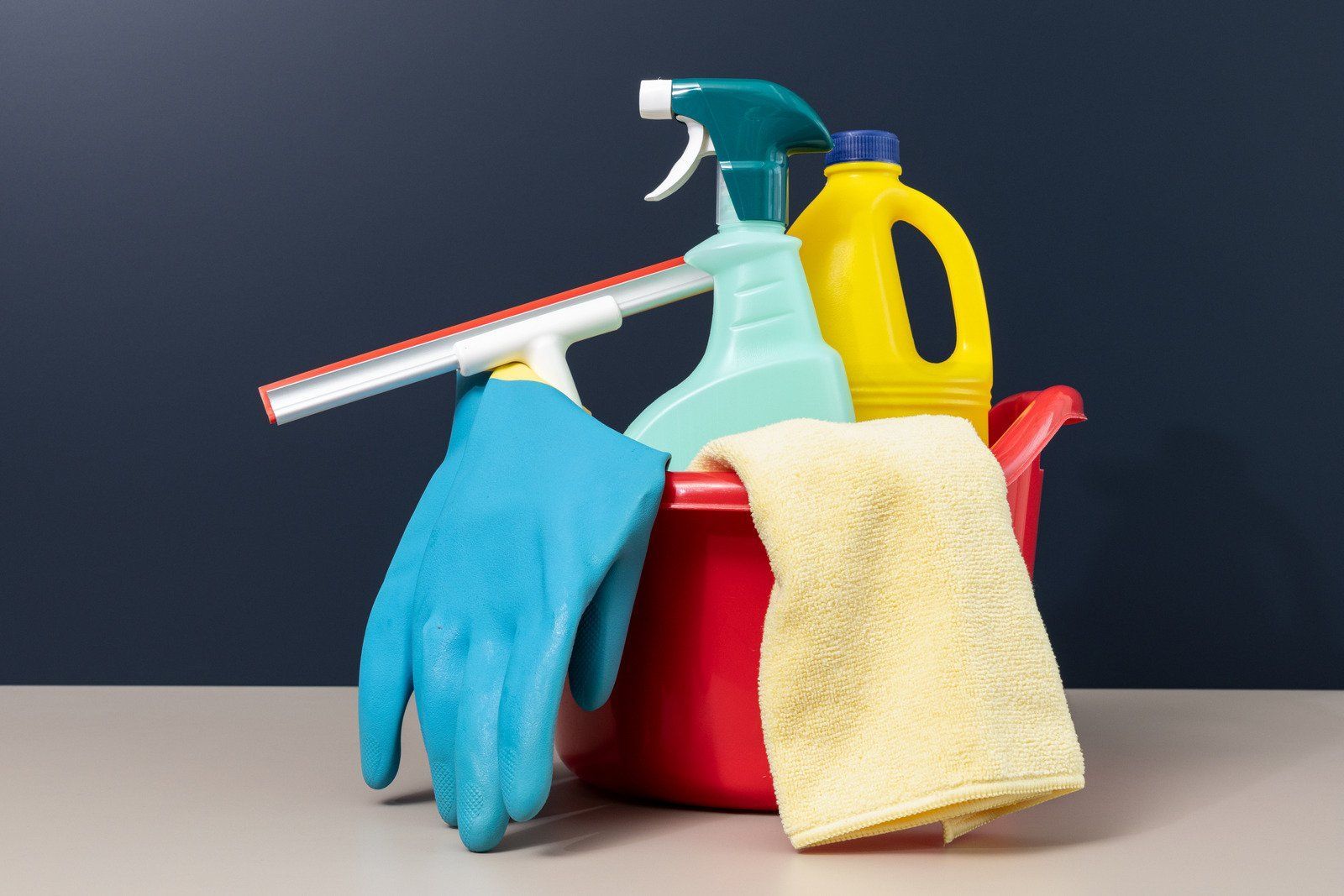 Cleaning supplies in a red bucket: spray bottles, gloves, squeegee, cleaning solution, and a yellow cloth.