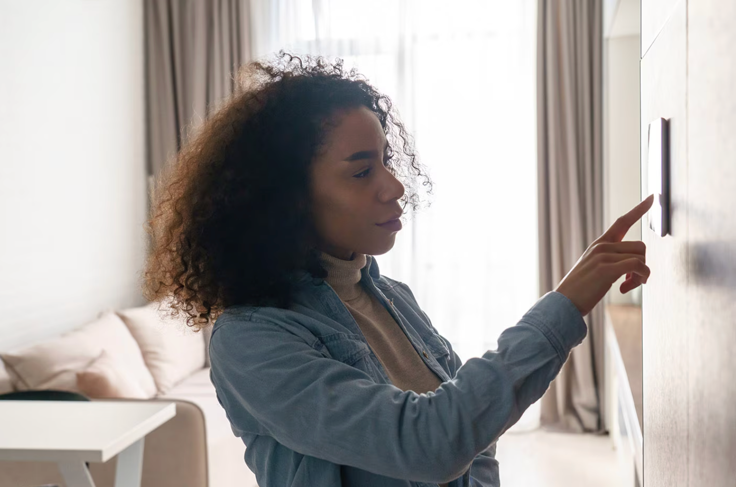 Woman with curly hair adjusting a smart home control panel on a wall in a living room.