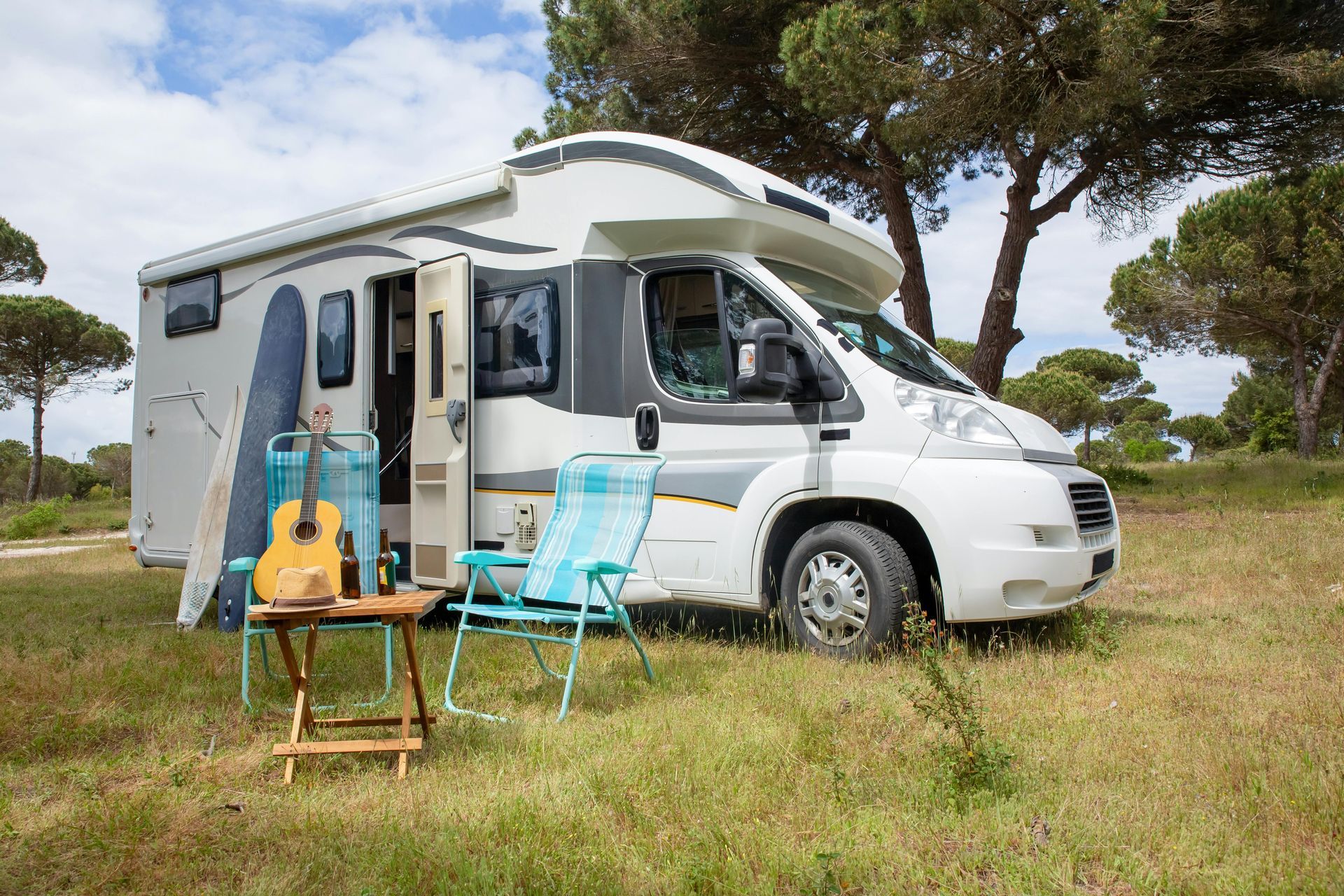 A rv is parked in a grassy field with a guitar , chairs and a surfboard.