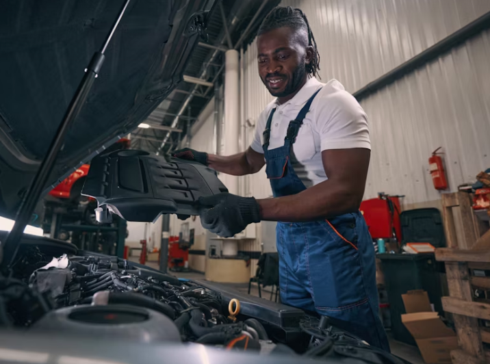 Mechanic in blue overalls inspecting a car engine with a black cover, smiling in a garage setting — D & B Plumbing NQ in Trinity Beach, QLD