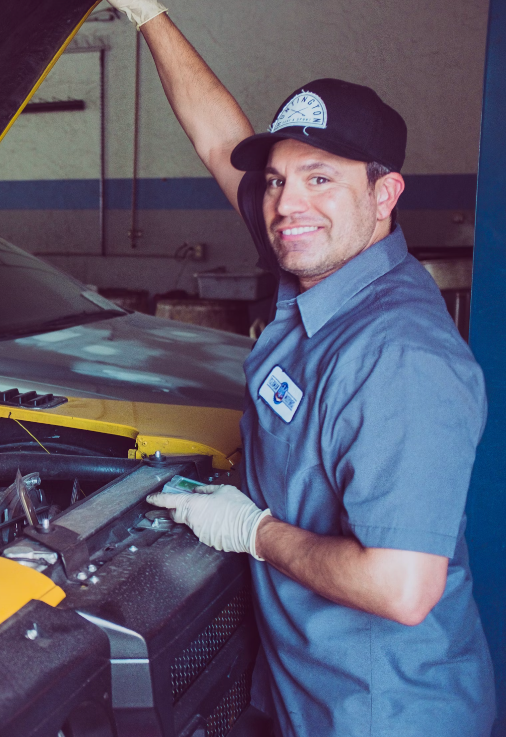 Mechanic smiling while working on a car in a garage, wearing a blue uniform and cap, with one arm raised under the car hood — D & B Plumbing NQ in Trinity Beach, QLD