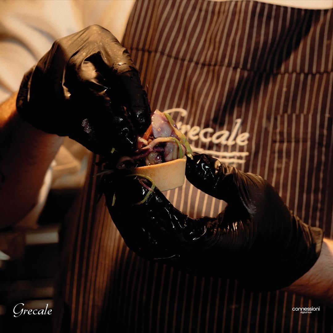 Una persona che indossa guanti neri e un grembiule con la scritta grecale