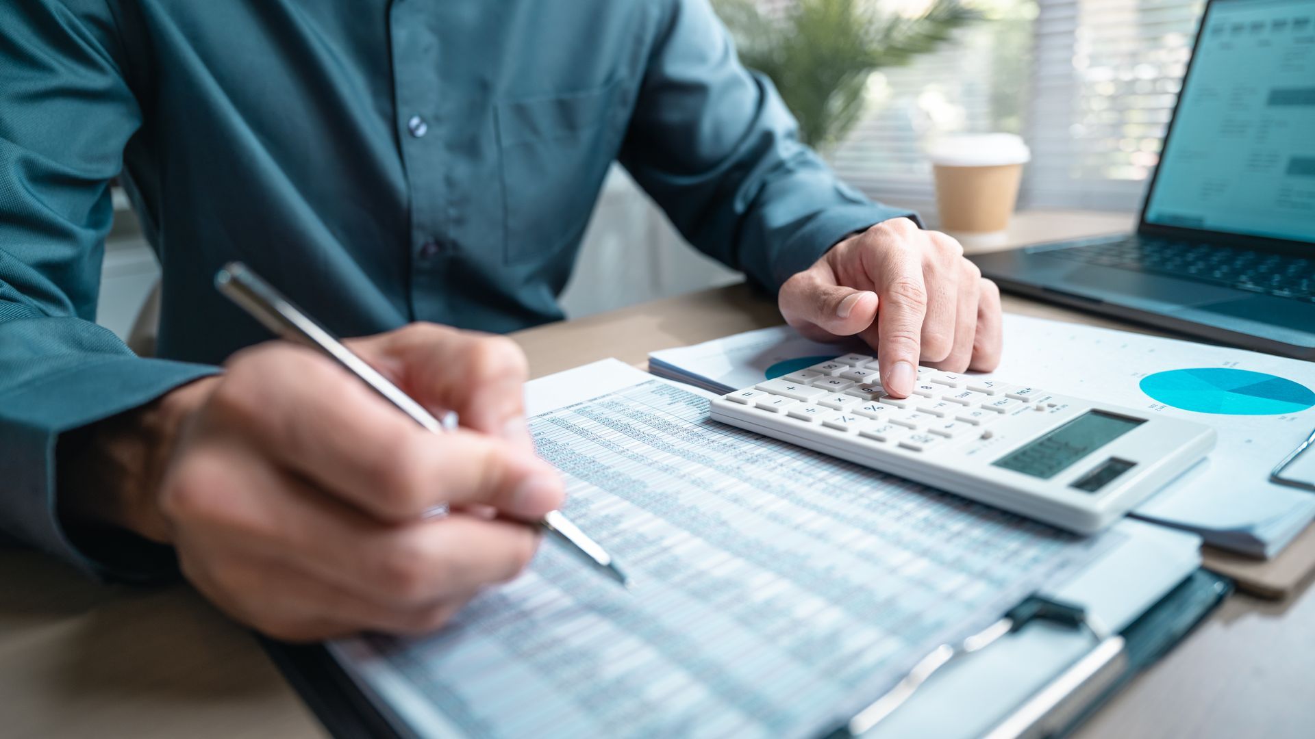 Person using a calculator and pen, working on financial documents at a desk with a laptop and coffee cup.