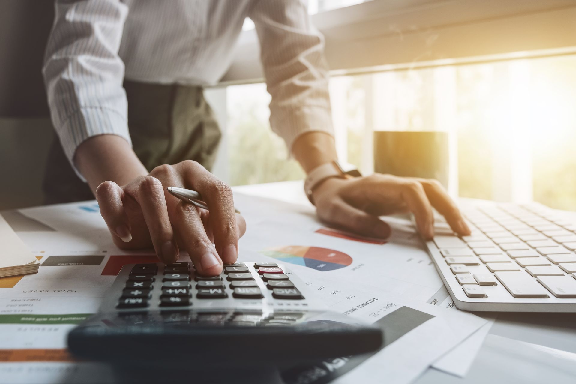 Person using a calculator and keyboard with financial documents on a desk, near a window. Person using a calculator and keyboard with financial documents on a desk, near a window.
