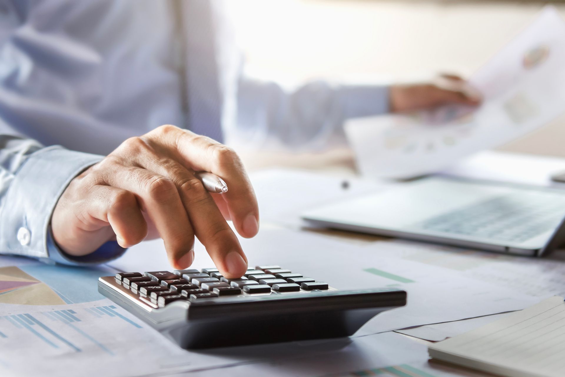Person's hand using a calculator; paperwork and laptop in background.