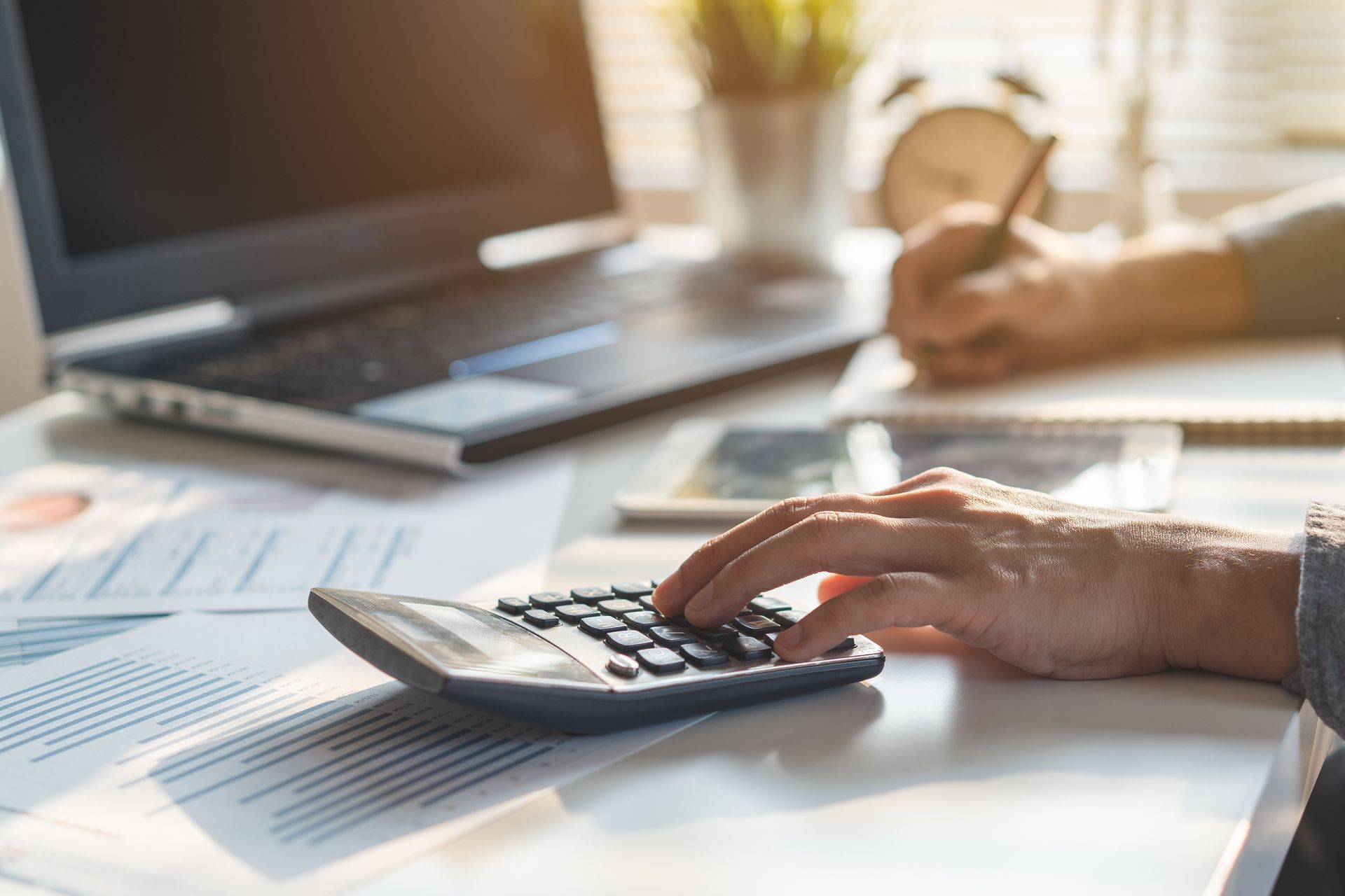 Person's hands using a calculator, laptop, documents, and writing on a desk; financial calculations.