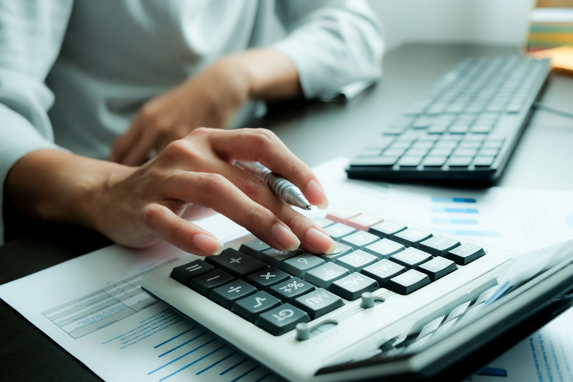 Person's hands using a calculator over documents and keyboard on a desk.
