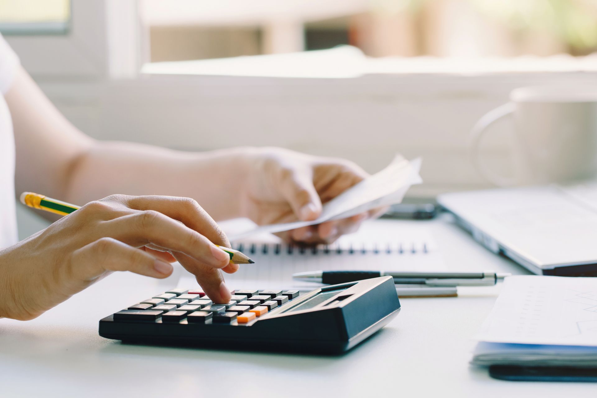 Person using a calculator, holding a receipt, and writing on a notepad at a desk.