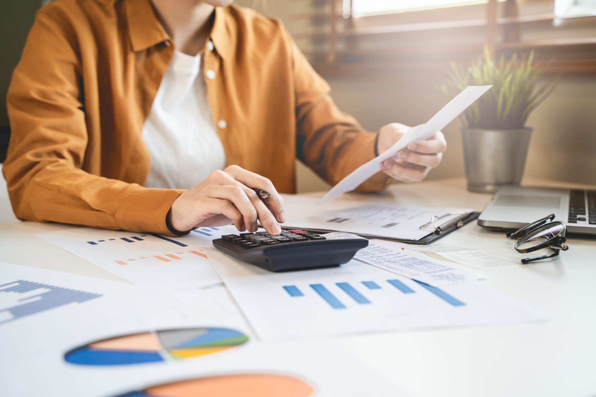 Person using a calculator, reviewing papers, and analyzing charts on a desk in an office setting.