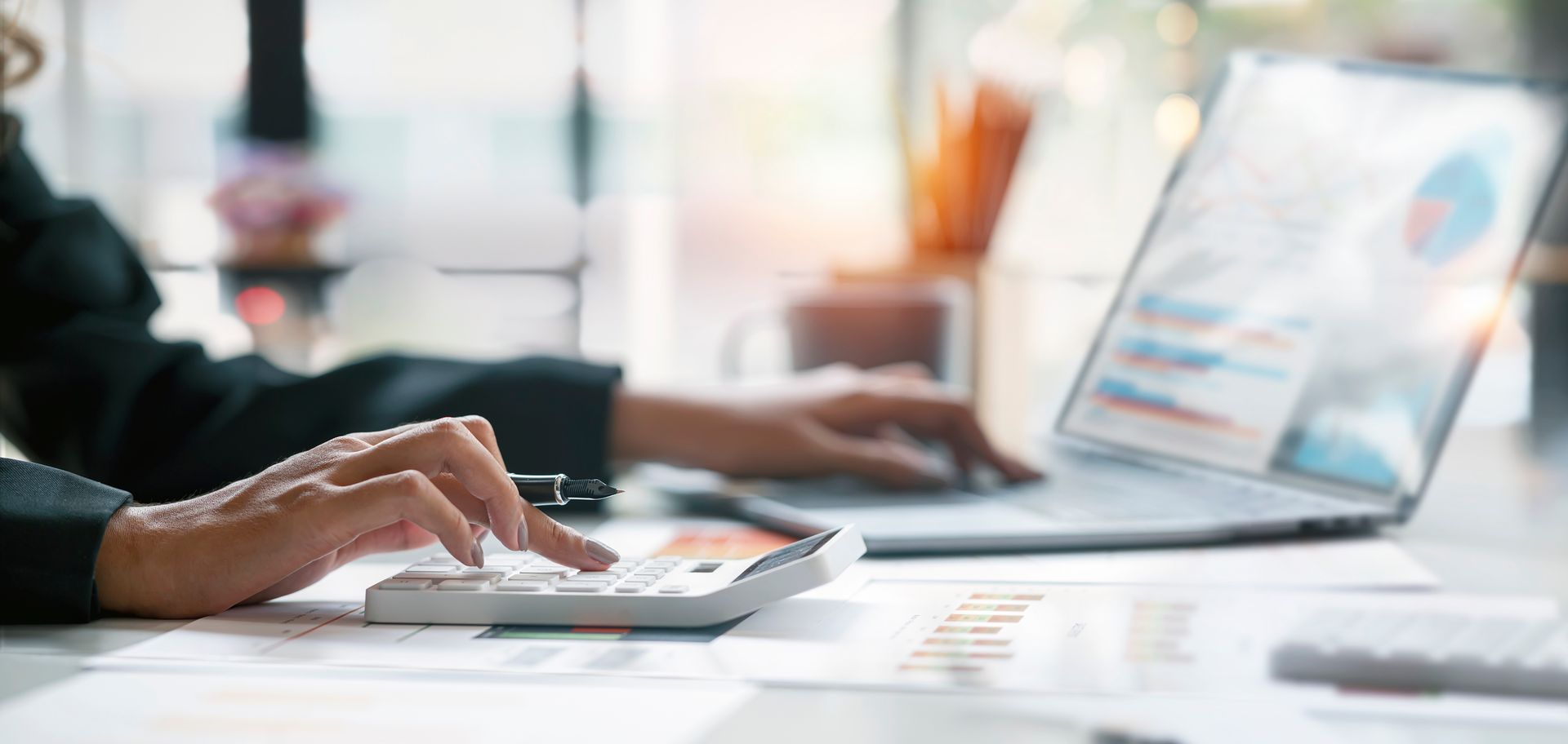 Person using a calculator and laptop, working on financial analysis, with charts and documents on a desk. Person using a calculator and laptop, working on financial analysis, with charts and documents on a desk.