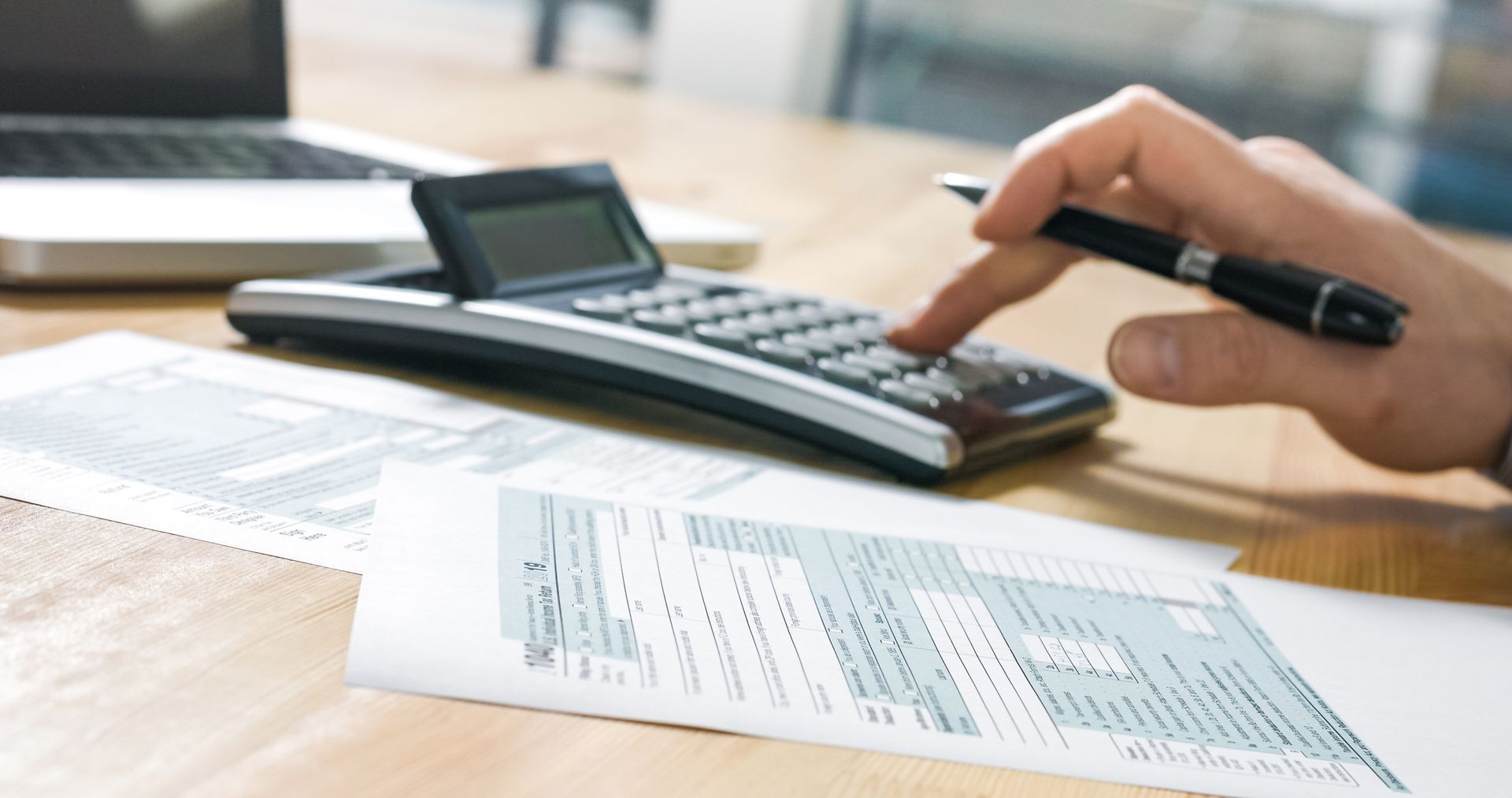Hand using calculator, with tax forms and laptop on a desk.