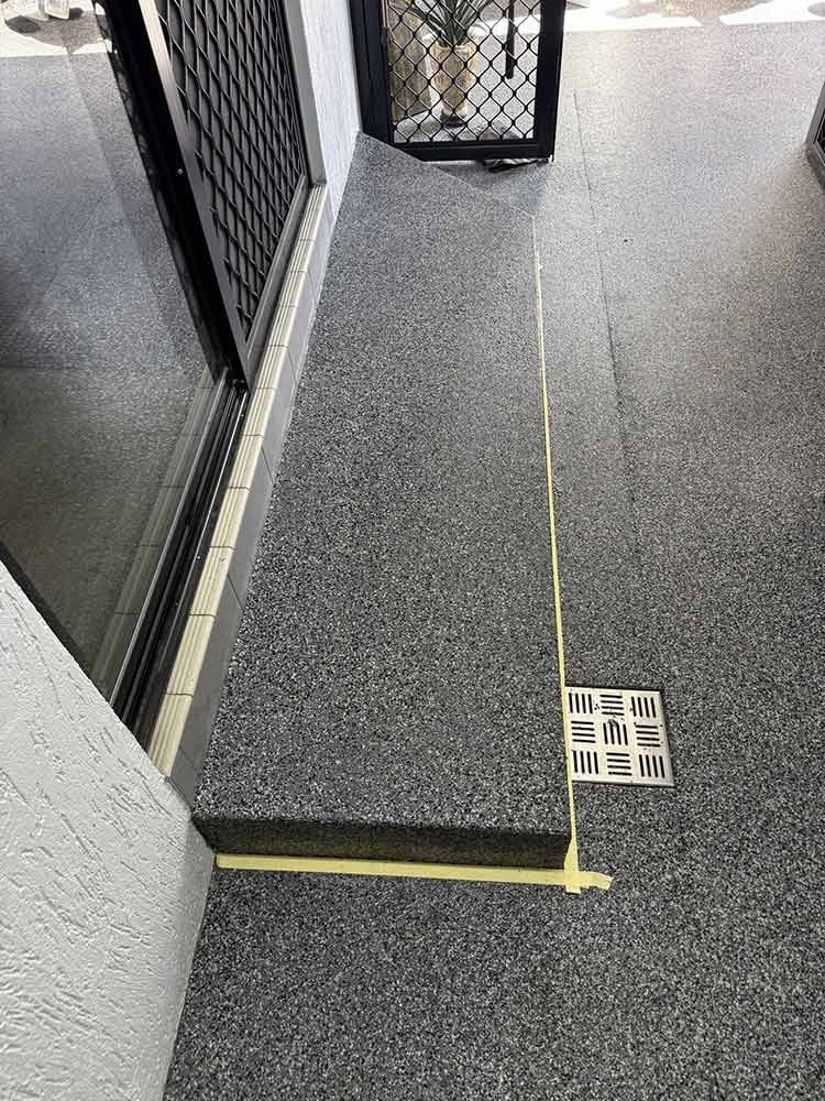 Dark speckled flooring with a drainage grate; adjacent to a wall, door, and window — All Grind & Coat in Cairns, QLD